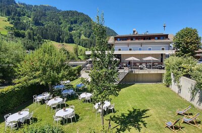 Außenbereich des Gasthof Auwirt mit gedeckten Tischen auf einer Wiese vor dem Gebäude und bewaldeten Bergen.