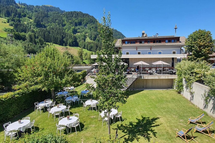 Außenbereich des Gasthof Auwirt mit gedeckten Tischen auf einer Wiese vor dem Gebäude und bewaldeten Bergen.