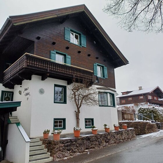 Haus Huber am Sonnenhofweg Zweistöckiges alpines Wohnhaus mit dunkler Holzfassade im Obergeschoss und weißem Putz im Erdgeschoss.