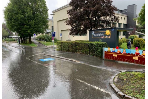 Behindertenparkplatz - Wagnerstraße 19 Ein Spielplatz mit einem Schild und bunten Figuren ist sichtbar. Die Straße ist nass vom Regen, und es gibt Bäume in der Umgebung.