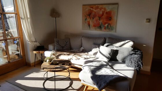 Wohnbereich Ferienwohnung Wielemans Helles Wohnzimmer mit grauem Ecksofa, rundem Holztisch und Blumenbild an der Wand.