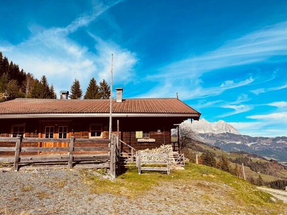 Rustikale Holzhütte mit Zaun vor einer Bergkulisse unter blauem Himmel.