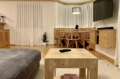 Wohnzimmer mit hellem Holztisch, grauem Sofa und einer Sitzecke aus Holz im Hintergrund.