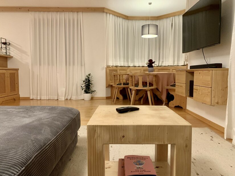 Wohnzimmer mit hellem Holztisch, grauem Sofa und einer Sitzecke aus Holz im Hintergrund.