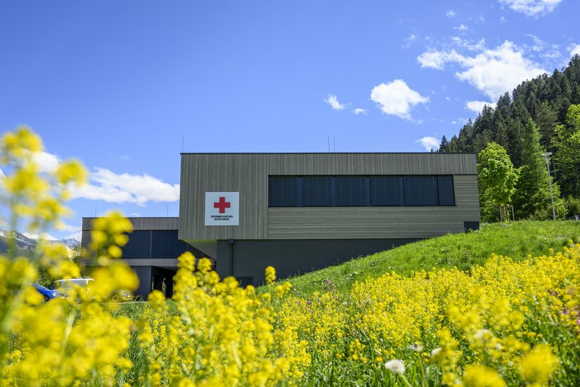 Modernes Gebäude des Österreichischen Roten Kreuzes hinter einer gelben Blumenwiese vor bewaldetem Hang.