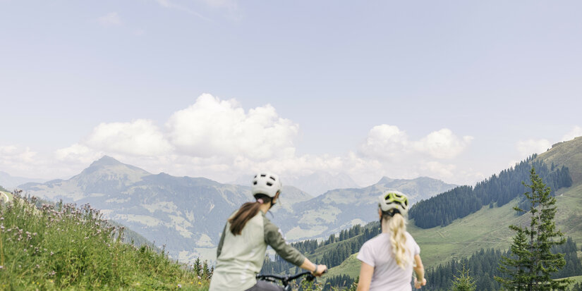 Zwei Mountainbikerinnen genießen bei den 360 Flow Trails in Kitzbühel die Aussicht auf die umliegenden Berge und das grüne Tal.
