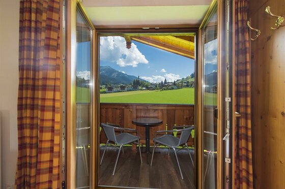 Blick durch eine offene Balkontür auf einen Holzbalkon mit zwei Stühlen und Aussicht auf eine Berglandschaft.