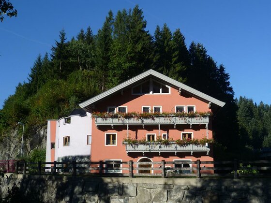 Mehrstöckiges Gasthaus mit rötlicher Fassade, Holzbalkonen und Blumenschmuck vor bewaldetem Hang.
