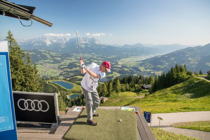 Ein Mann beim Golfabschlag auf einer hölzernen Plattform vor einem weiten Bergpanorama.