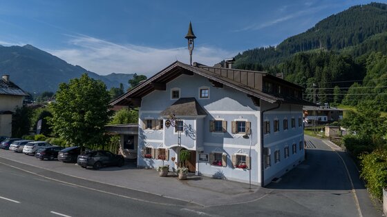 Gasthof-Auwirth_Außenaufnahmen_16062025_(c)_Floobe Ein charmantes Gebäude im alpinen Stil mit blauen Wänden und Fensterläden. Umgeben von Bergen und grüner Natur, bietet es eine einladende Atmosphäre.