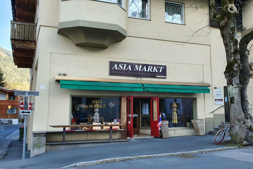 Asia Markt <p>Ein asiatischer Markt mit einem Schaufenster und einer rot lackierten Tür. Vor dem Geschäft stehen ein Fahrrad und einige Produkte sind ausgestellt.</p>