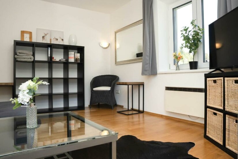 Ein modernes Wohnzimmer mit einem gläsernen Tisch, einem schwarzen Regal und einem Sofa. Große Fenster lassen viel Licht herein und sorgen für eine einladende Atmosphäre.