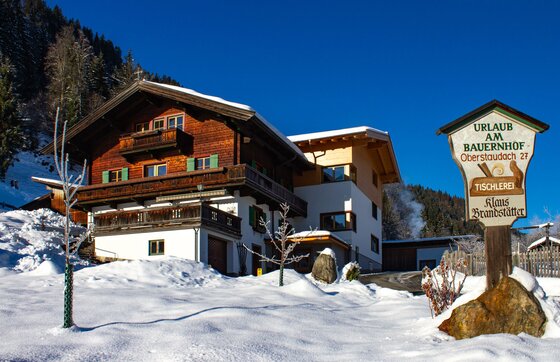 Ferienhof Oberstaudach im Winter Ein traditionelles Bauernhaus aus Holz und Stein in einer verschneiten Winterlandschaft unter blauem Himmel.