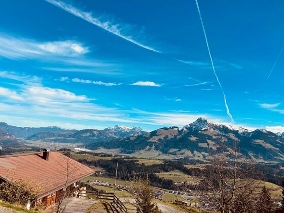 Blick von einer Almhütte auf ein weites Tal und schneebedeckte Berggipfel unter blauem Himmel.