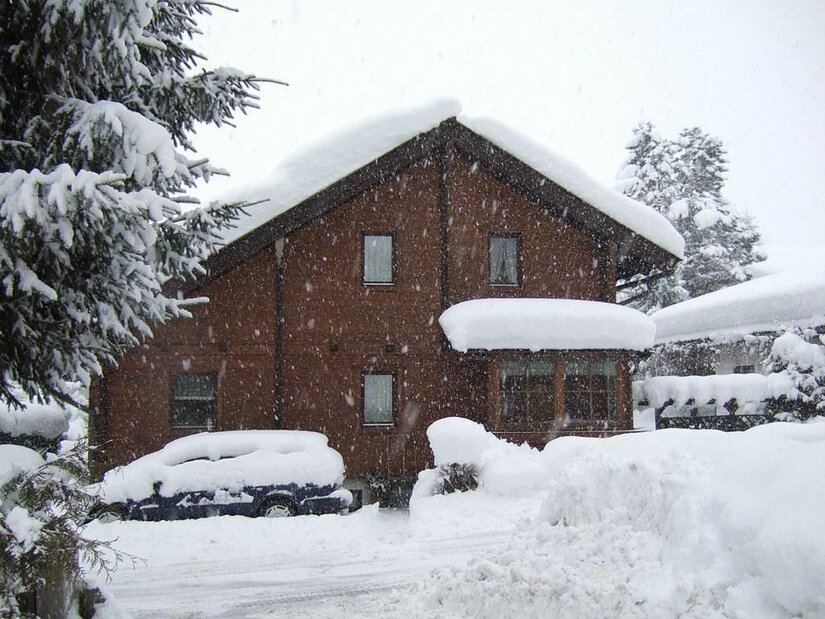 Ein braunes Holzhaus mit schneebedecktem Dach und ein eingeschneites Auto bei starkem Schneefall.
