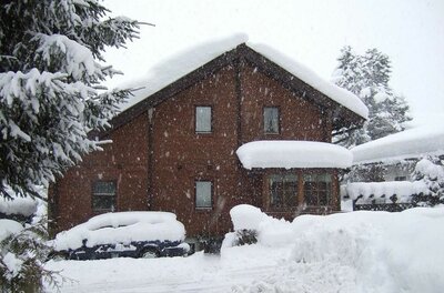 Haus Pirchner im Winter Ein braunes Holzhaus mit schneebedecktem Dach und ein eingeschneites Auto bei starkem Schneefall.