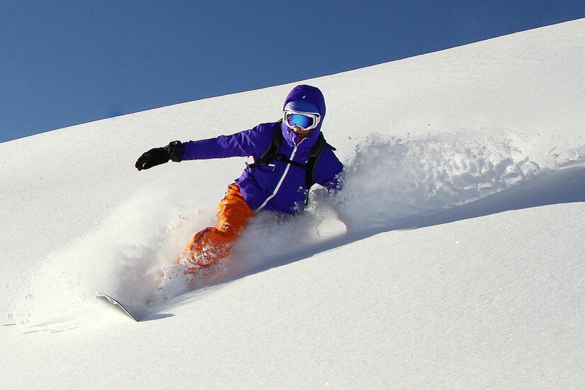 Snowboarden im Tiefschnee Eine Person in violetter Jacke und orangefarbener Hose fährt Snowboard im Tiefschnee.