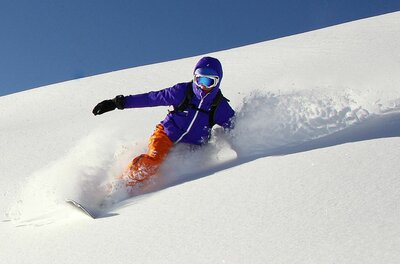 Eine Person in violetter Jacke und orangefarbener Hose fährt Snowboard im Tiefschnee.