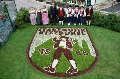 Eine Gruppe von Menschen in traditionellen Kleidern steht vor einem kunstvoll angelegten Blumenbeet in Form eines Wappens. Das Wappen zeigt einen Musiker und die Jahreszahlen 1866 und 2004.