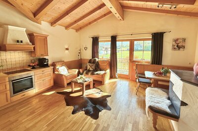 IMG_6582 Ein gemütliches Wohnzimmer mit Holzdecke und einem großen Fenster. Die Einrichtung besteht aus bequemen Sesseln, einem Tisch und einer modernen Küche.