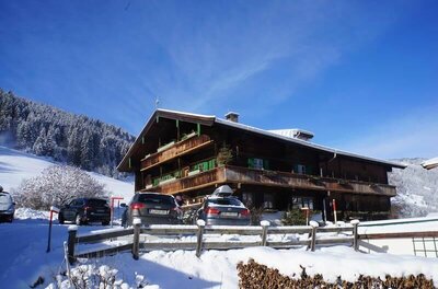 MyApart 1709 Jochberg im Winter Großes Holzhaus im alpinen Stil mit Balkonen und parkenden Autos in einer verschneiten Winterlandschaft.