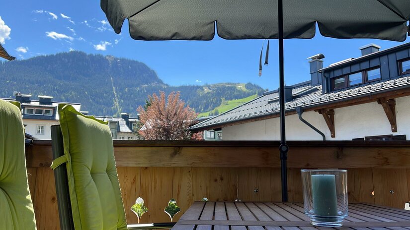 Balkon mit Holztisch, grünem Stuhlpolster und Sonnenschirm vor bewaldeter Bergkulisse.