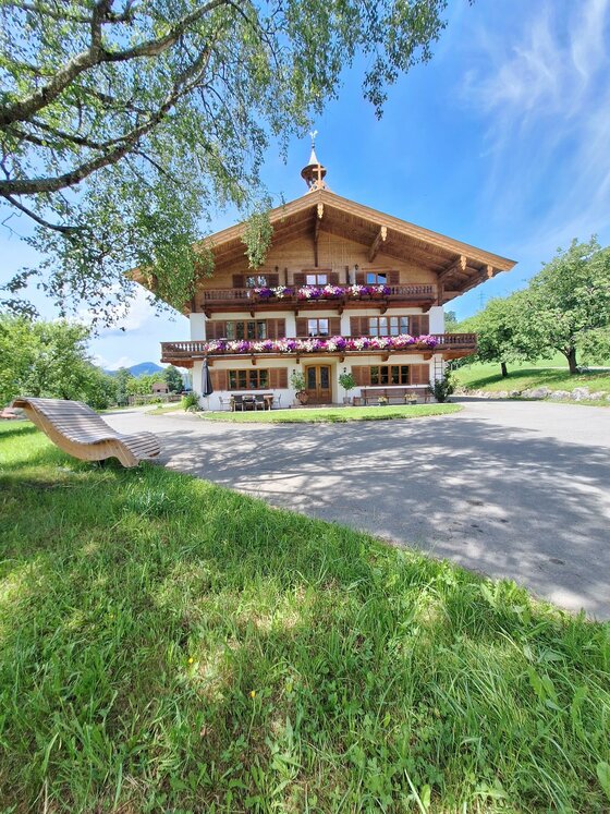 20260608_135437 Ein traditionelles alpenländisches Holzhaus mit Balkonen und bunten Blumen. Umgeben von grünen Wiesen und unter einem klaren blauen Himmel.