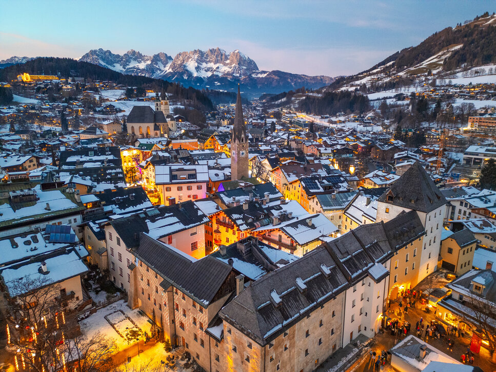 Die malerische Stadt Kitzbühel erstrahlt in festlichem Licht, umgeben von schneebedeckten Bergen und historischen Gebäuden.