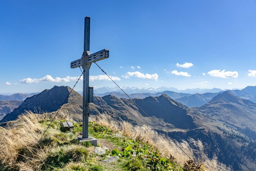 Ein metallisches Gipfelkreuz auf einem grasbewachsenen Berggipfel vor einem Panorama aus weiteren Gebirgsketten.
