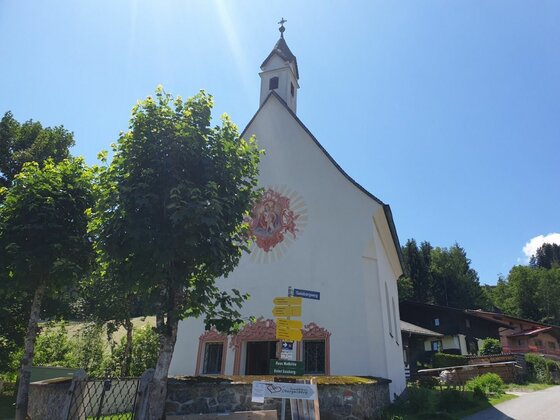 Kapelle am Gaisberg Weiße Kapelle mit Wandmalerei und gelben Wegweisern im Vordergrund unter blauem Himmel.