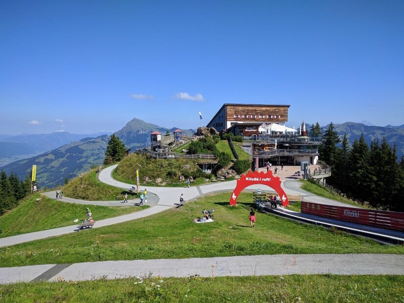Berghaus am Hahnenkamm Berghaus auf einem Gipfel mit befestigten Wanderwegen und einem roten Torbogen unter blauem Himmel.