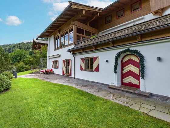 Haus Schmidt in Jochberg Alpines Wohnhaus mit weißer Fassade, rot-weiß gestreiften Fensterläden und einer verzierten Holztür vor einer Rasenfläche.