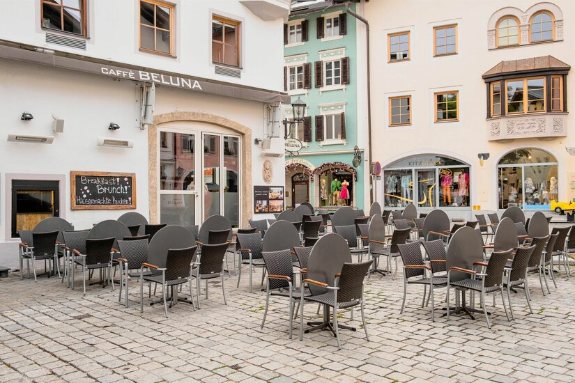 Außenbereich eines Cafés mit Tischen und Stühlen auf einem gepflasterten Platz vor historischen Gebäuden.