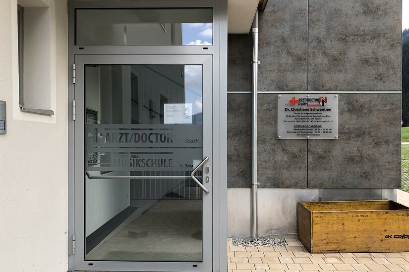 Eingangsbereich einer Arztpraxis mit Glastür und Hinweisschild an der Fassade.