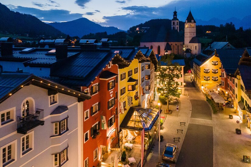 Beleuchtete Häuserzeile in einer gepflasterten Altstadt bei Dämmerung mit Bergen im Hintergrund.
