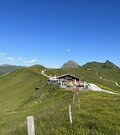 Berghütte mit Terrasse auf einem grünen Bergrücken vor alpinem Panorama unter blauem Himmel.