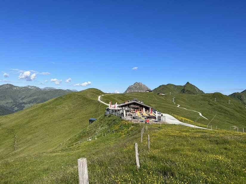 Berghütte mit Terrasse auf einem grünen Bergrücken vor alpinem Panorama unter blauem Himmel.