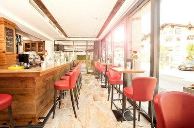 Innenansicht der Reisch Bar Moderner Barbereich mit Holztresen, roten Barhockern und großen Glasfronten.