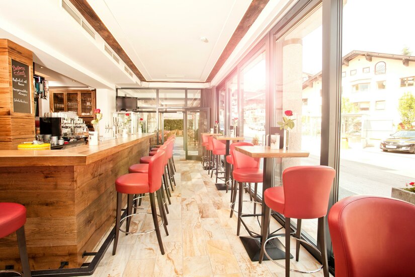 Innenansicht der Reisch Bar Moderner Barbereich mit Holztresen, roten Barhockern und großen Glasfronten.