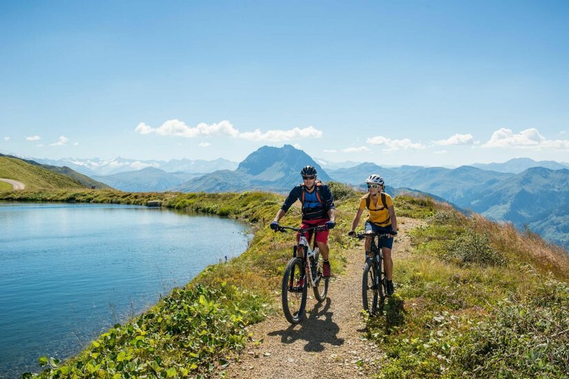 Zwei Mountainbiker fahren auf einem Schotterweg an einem Bergsee vorbei.