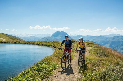 Mountainbiken am Bergsee Zwei Mountainbiker fahren auf einem Schotterweg an einem Bergsee vorbei.