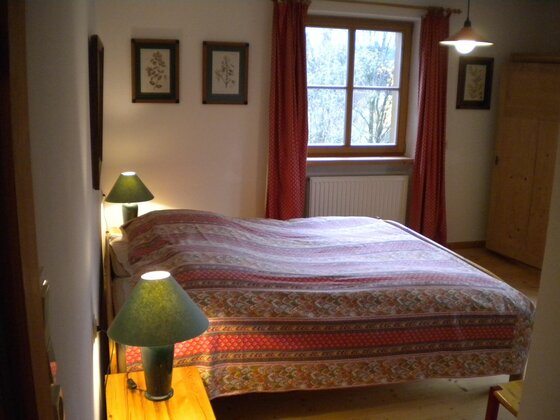 Schlafzimmer 1 mit Bad Schlafzimmer mit Doppelbett, zwei Nachttischlampen, Fenster mit roten Vorhängen und Holzschrank.