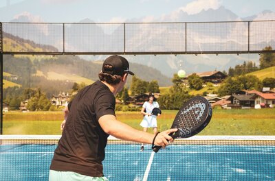 Eine Frau und ein Mann spielen Padeltennis auf dem blauen Platz. Zwischen ihnen ist das Netz, im Hintergrund sieht man Häuser und den Wilden Kaiser.