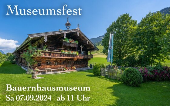 Historisches Bauernhaus aus dunklem Holz mit Blumenschmuck auf einer grünen Wiese unter blauem Himmel.