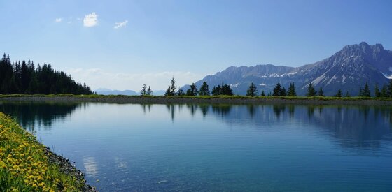 Blick auf den Bergsee Ein klarer Bergsee mit Spiegelung der Bäume vor einem Gebirgsmassiv unter blauem Himmel.