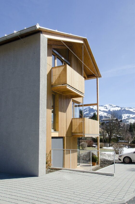 Modernes Gebäude mit Holzfassade, Balkonen und Blick auf verschneite Berge unter blauem Himmel.