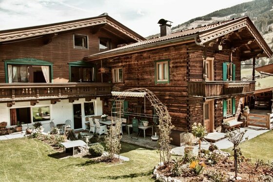 Zweistöckiges Holzhaus im alpinen Stil mit Garten, Terrasse und Balkon vor bewaldetem Hang.