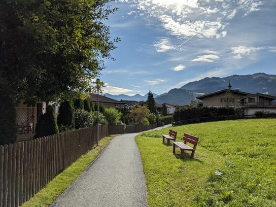 Asphaltierter Spazierweg mit zwei Holzbänken neben einer Wiese und Wohnhäusern vor Gebirgskulisse.