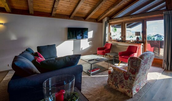 Salon RÉSIdence Reith Kitzbühel Wohnzimmer mit dunklem Sofa, roten Sesseln, Couchtisch und Fernseher unter einer Holzdecke.