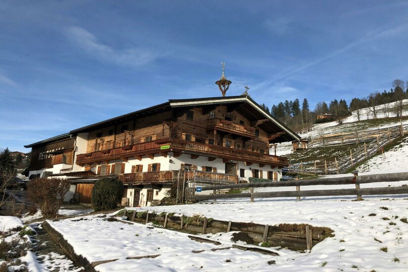 Traditioneller Bauernhof aus Holz und weißem Putz in einer verschneiten Hanglage unter blauem Himmel.